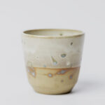 ceramiczny kubeczek z beżowej gliny szkliwiony szaro-piaskowym szkliwem pochlapany na grafitowo z niebiesko-zielonymi przebarwieniami