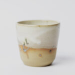 ceramiczny kubeczek z beżowej gliny szkliwiony szaro-piaskowym szkliwem pochlapany na grafitowo z niebiesko-zielonymi przebarwieniami