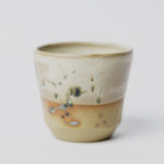 ceramiczny kubeczek z beżowej gliny szkliwiony szaro-piaskowym szkliwem pochlapany na grafitowo z niebiesko-zielonymi przebarwieniami