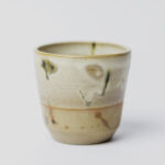 ceramiczny kubeczek z beżowej gliny szkliwiony szaro-piaskowym szkliwem pochlapany na grafitowo z niebiesko-zielonymi przebarwieniami
