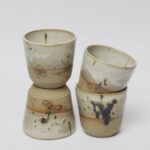 ceramiczne kubeczki z beżowej gliny szkliwione szaro-piaskowym szkliwem pochlapane na grafitowo z niebiesko-zielonymi przebarwieniami